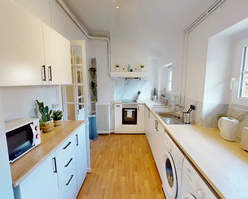 Cucina a corridoio luminosa e moderna con mobili bianchi, piani in legno, forno integrato e lavatrice. Stile pulito e minimale con piccole piante e luce naturale dalle finestre.