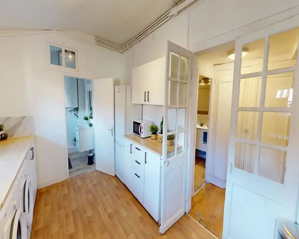 Angolo cucina luminoso e moderno con pensili bianchi, piano di lavoro in legno, microonde e frigorifero alto; pavimento in legno chiaro e piccole piante decorative creano un ambiente ordinato e accogliente.
