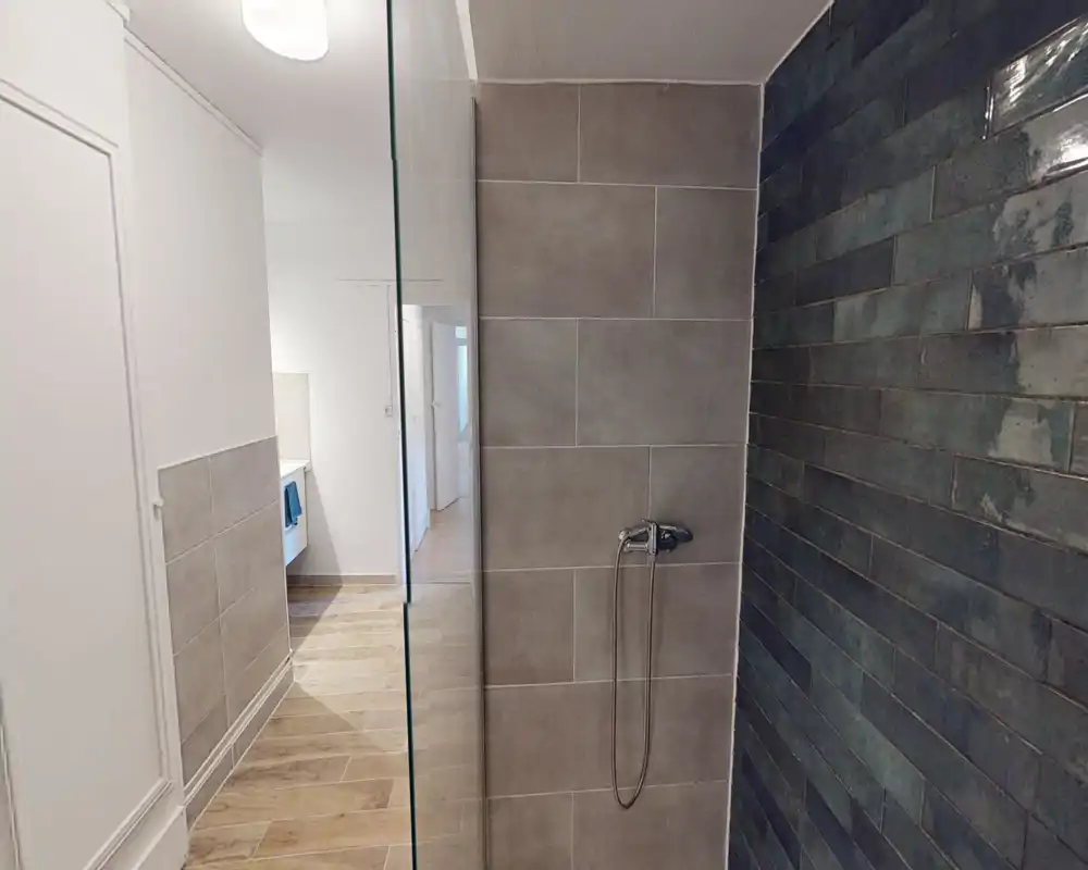 Doccia walk-in moderna con piastrelle grigie formato grande sulla parete centrale e piastrelle blu-verdi a listelli sulla parete laterale; miscelatore a mano e pannello in vetro visibili.