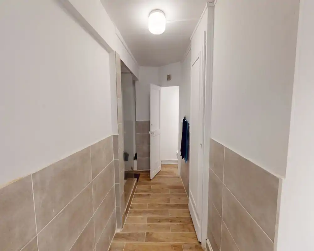 Ingresso/corridoio stretto con piastrelle sulle pareti inferiori e pavimento effetto legno, luce a soffitto e porta verso una stanza adiacente.