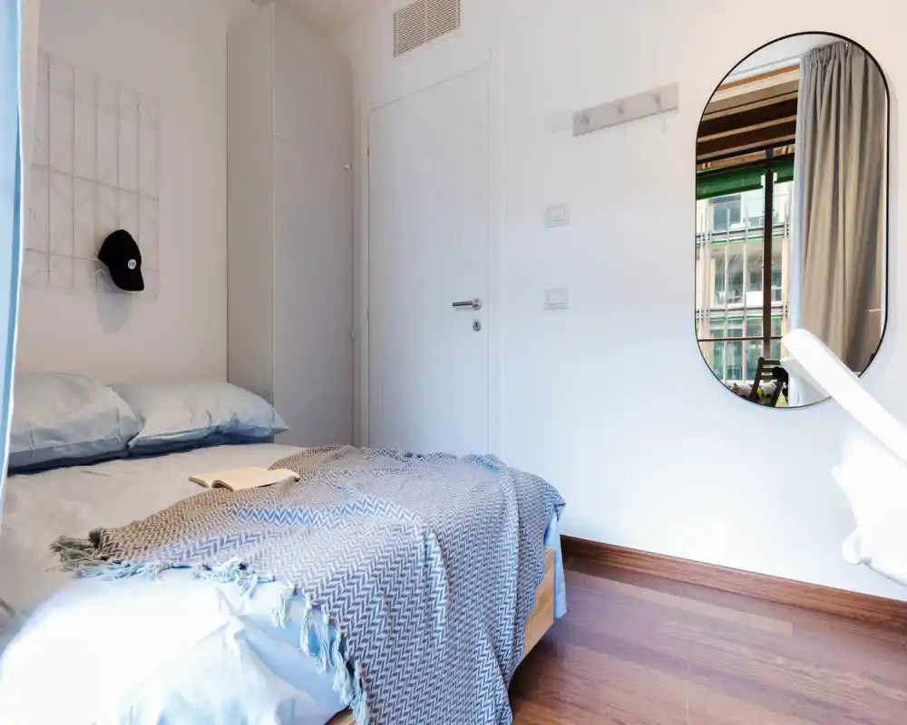 Bright, compact bedroom with a neatly made bed, textured throw and a wall mirror that adds depth. Natural light from the window highlights wooden floors and simple, modern furnishings.