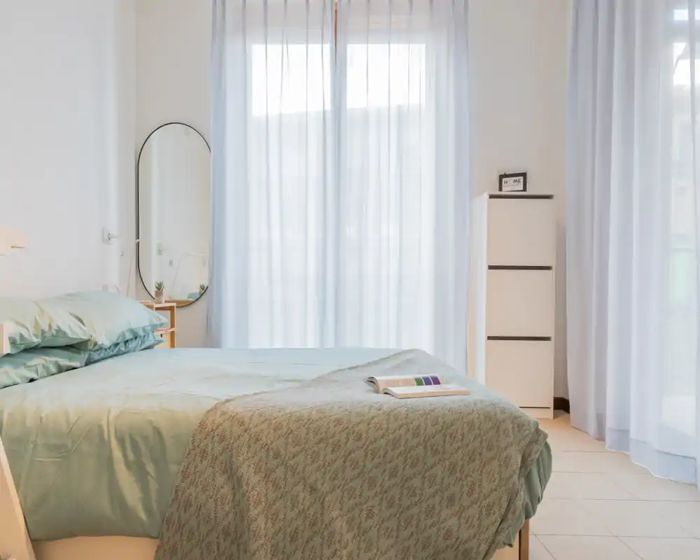 Bright, airy bedroom with a neatly made double bed, soft teal linens and a textured throw. Sheer curtains filter abundant natural light and a tall narrow dresser plus a mirror complete the minimalist, cozy setup.