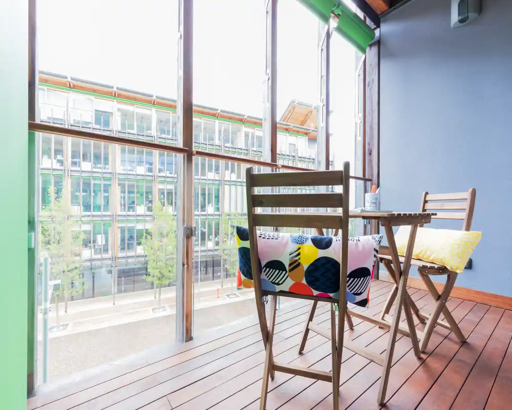 Sunny furnished balcony with wooden decking, a small folding table and chairs, and colorful cushions — ideal for morning coffee with a street view.