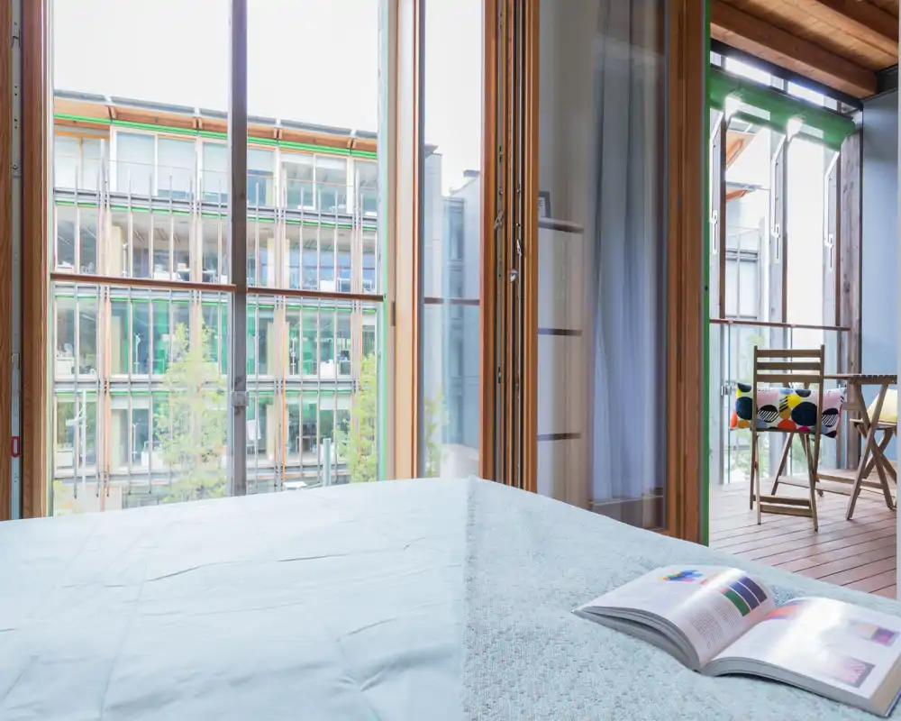 Bright, modern bedroom with a large bed in the foreground and floor-to-ceiling windows opening to a small wooden balcony. Light textiles, an open book on the bed and outdoor seating create a welcoming, airy atmosphere.