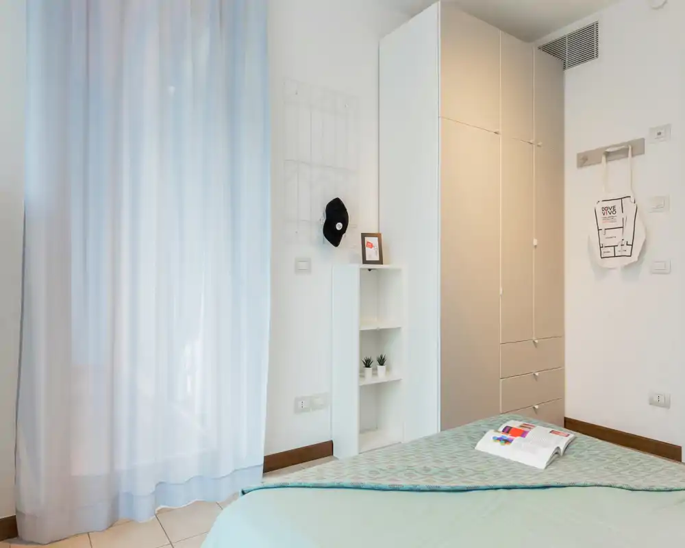 Bright, minimal bedroom with a neatly made bed, tall wardrobe and soft natural light from a curtained window — fresh, calm styling ideal for rental listings.