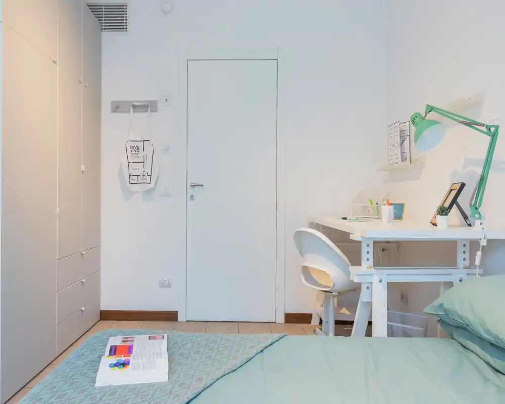 Bright, minimalist bedroom with a visible bed in the foreground, a tidy workspace with a white desk and green lamp, and built-in storage along the left wall.