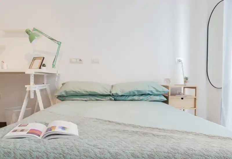 Dormitorio luminoso y minimalista con cama doble bien hecha, ropa de cama verde menta, mesita con lámpara y espejo en la pared. Composición aireada y estilizada para un anuncio de alquiler.