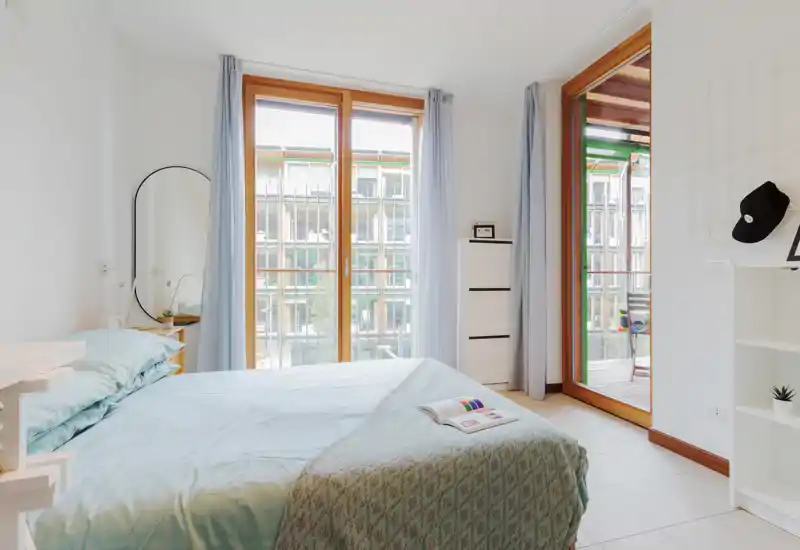 Dormitorio moderno y luminoso con grandes ventanas y cama bien hecha; textiles claros y decoración minimalista crean un ambiente tranquilo y aireado.