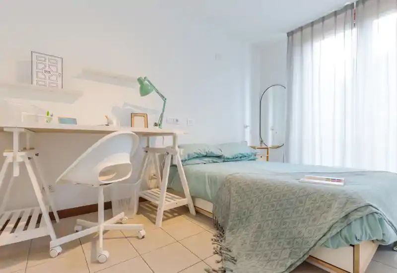 Dormitorio luminoso y bien decorado con cama completa, ropa de cama en tono teal y una zona de trabajo. Gran ventana con cortinas translúcidas que aporta abundante luz natural y una atmósfera moderna y tranquila.