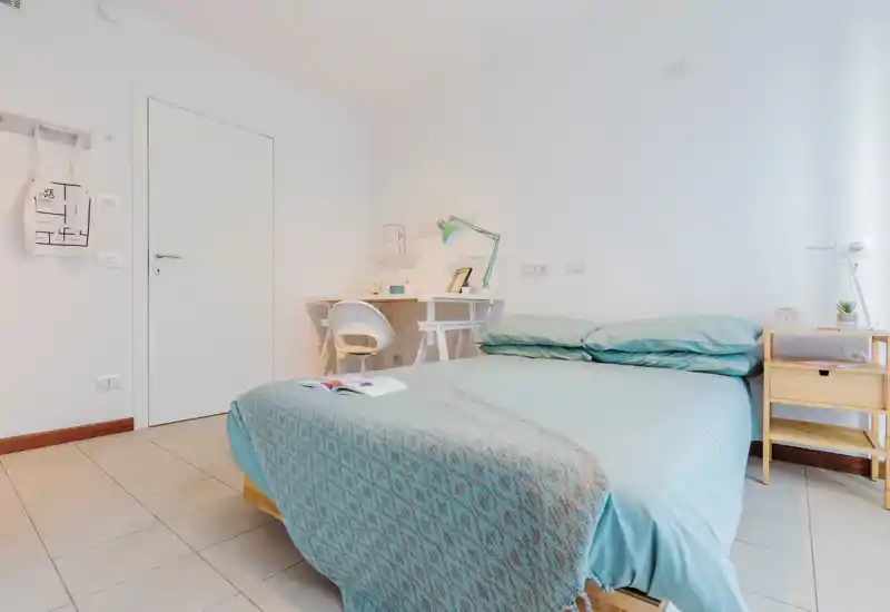 Dormitorio luminoso y bien preparado con cama doble vestida en tonos verde azulado, mesita de noche y un escritorio compacto con silla — ideal para una estancia cómoda.