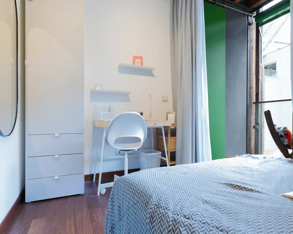 Bright, modern bedroom with a comfortable double bed in the foreground, a small work desk and chair against the wall, and a wardrobe. Natural light from a large opening and soft neutral tones create an airy, calm atmosphere ideal for short-term rentals.