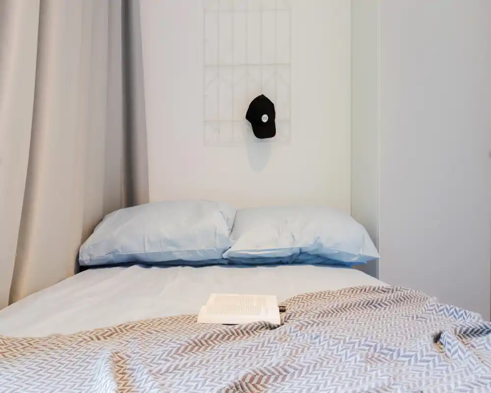 Bright, minimal bedroom with a neatly made bed, soft blue pillows and a patterned blanket; a book lies open on the bed for a lived-in touch.