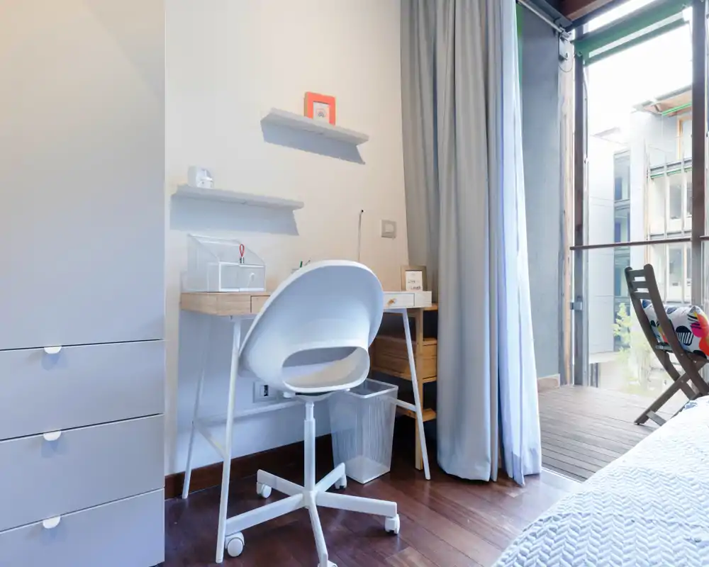 Bright, modern bedroom corner with a compact desk and chair beside a partially visible bed and a balcony. Natural light fills the space and the tidy desk area is ideal for work or study.