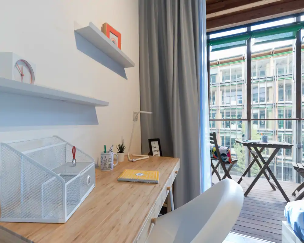 Compact bright bedroom workspace with a wooden desk, modern chair and balcony access that fills the room with natural light.