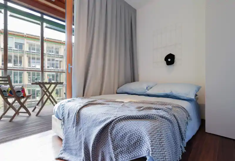 Bright, cozy bedroom with a neatly made double bed, soft blue linens and a patterned throw. Large glass doors open to a small balcony with folding chairs, flooding the room with natural light.
