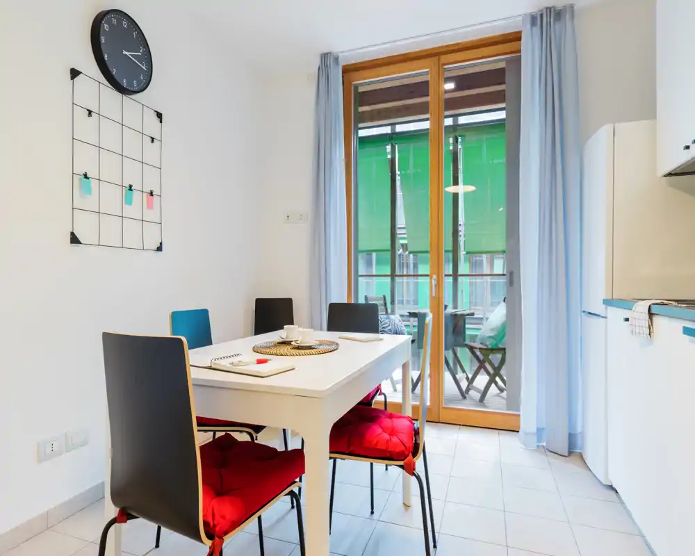 Bright, modern dining area with a white table, four chairs with red cushions, and a sliding door opening to a small balcony. Clean minimalist styling and natural light make it inviting for meals.