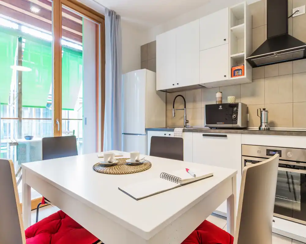 Modern, bright kitchen with a white dining table set for two, built-in oven, microwave and fridge. Large glass door provides natural light and access to a small balcony.
