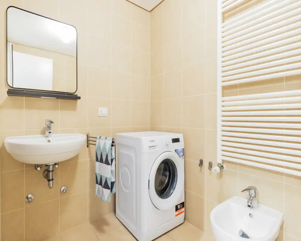Bright, modern bathroom with a wall-mounted sink, bidet, towel rail and a front-loading washing machine; light beige tiles and a mirrored cabinet provide a clean, functional space.