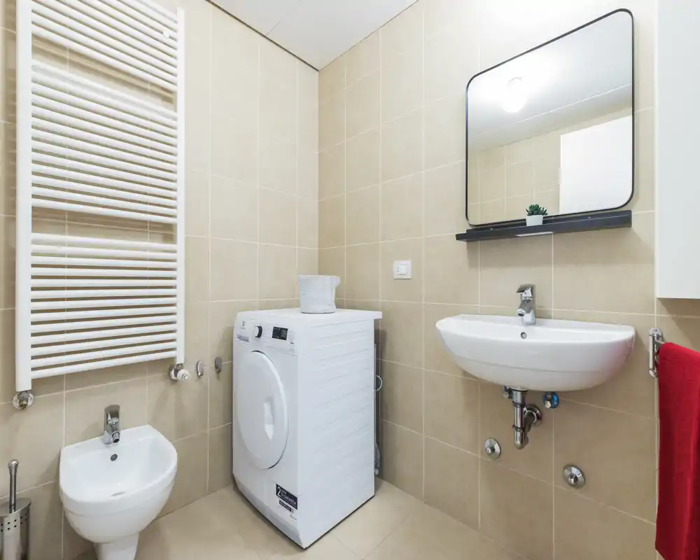 Compact, modern bathroom with beige tiles, wall-mounted sink, bidet, and a washing machine. Clean styling and bright lighting make this bathroom functional for small apartments.