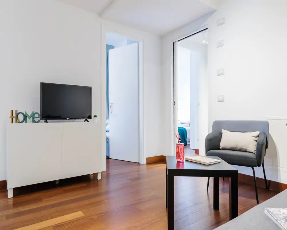 Bright, modern living area with a comfortable armchair, low coffee table and TV cabinet. Warm wooden flooring and clean white walls create a fresh, welcoming atmosphere.