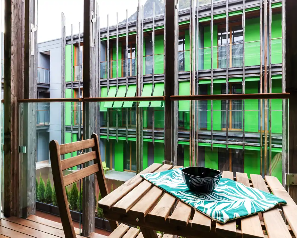 Small wooden balcony with a folding table and chair overlooking a modern courtyard with vivid green façades; a patterned cloth and a black bowl sit on the table.