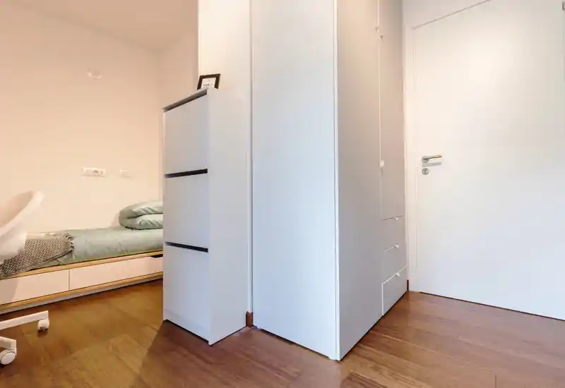Neat small bedroom with a single bed pushed to the wall, light wooden floor and white storage units. The room features tidy bedding and a minimalist wardrobe, ideal for a compact rental.