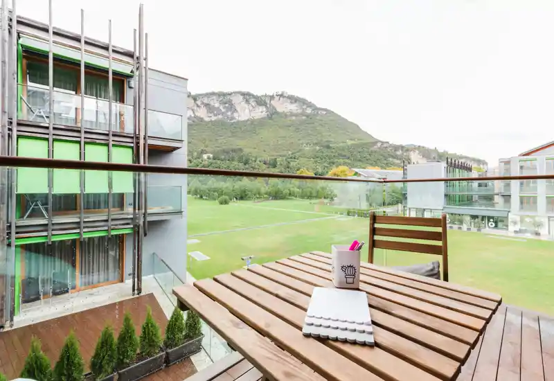 Sunny balcony with wooden table and chair, glass railing and a wide green lawn leading to scenic hills—ideal for relaxing or working outdoors.
