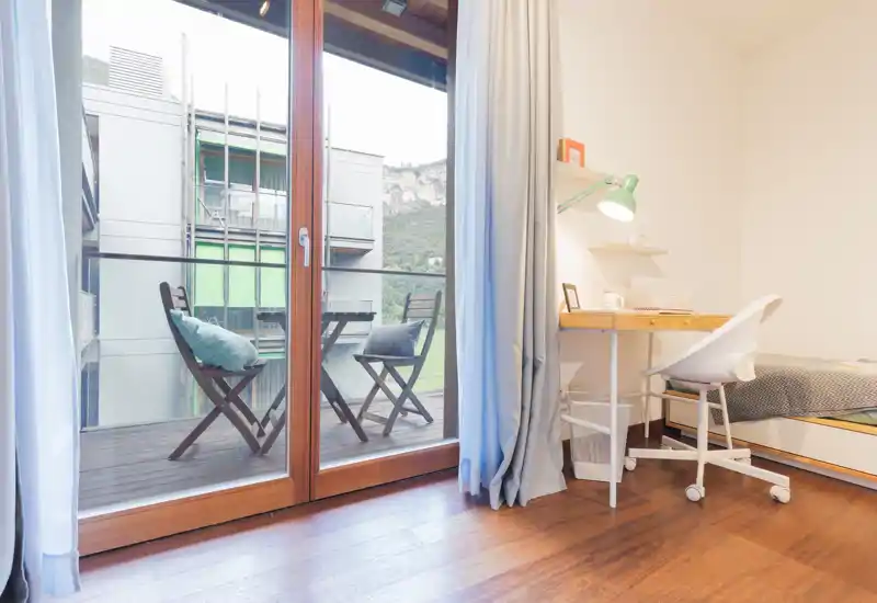 Bright, modern bedroom with a single bed, a small desk and a large glass door opening to a balcony with outdoor seating — ideal for natural light and a quiet workspace.