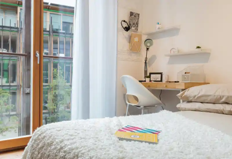 Bright, cozy bedroom corner with a plush white throw on the bed, neutral pillows and a small wooden desk by a large window letting in abundant natural light.