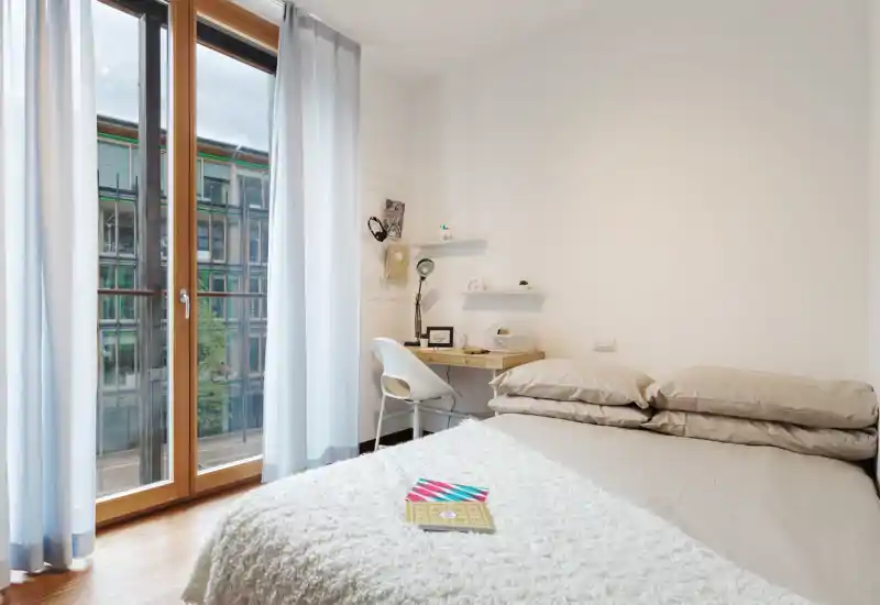 Bright, minimal bedroom with a large balcony window. Neutral bedding, a cozy throw and a small desk create a clean, livable space ideal for rentals.