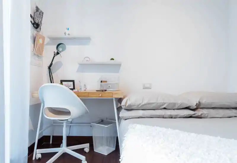 Bright, minimalist bedroom with a comfortable bed layered with neutral pillows and a small wooden desk with a lamp and chair — ideal for work-and-rest balance.