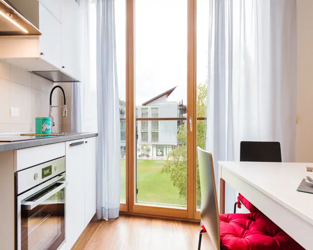 Bright modern kitchenette with built-in oven, sink and large floor-to-ceiling window overlooking green space; simple dining area with a colorful seat cushion.