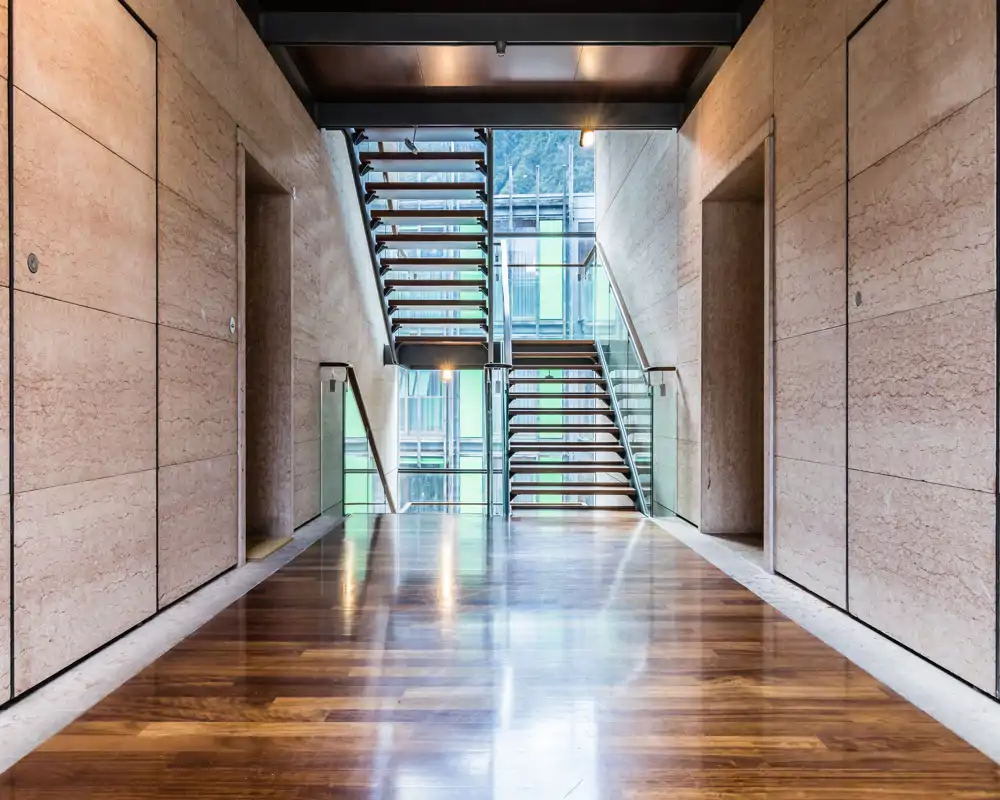 Modern building entrance or lobby with a wide polished wooden floor and a central glass-and-steel staircase leading up. The space feels clean, minimal and well lit, ideal for an apartment or office entry area.
