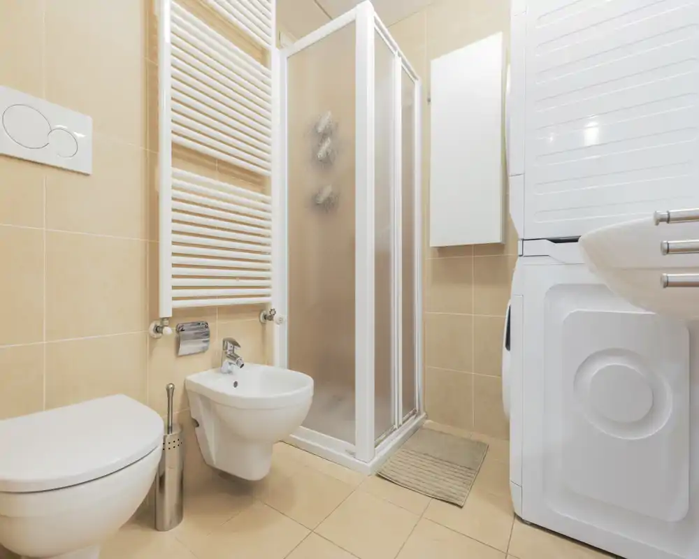 Modern, compact bathroom with wall-mounted toilet and bidet, a corner shower cubicle, towel radiator and stacked washer/dryer. Neutral beige tiles and bright lighting create a clean, functional space ideal for an apartment.