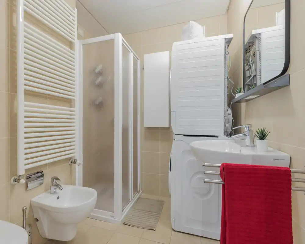 Compact, well-lit bathroom with a shower cubicle, wall-mounted bidet, sink with mirror and stacked washer-dryer unit — practical layout for small spaces.