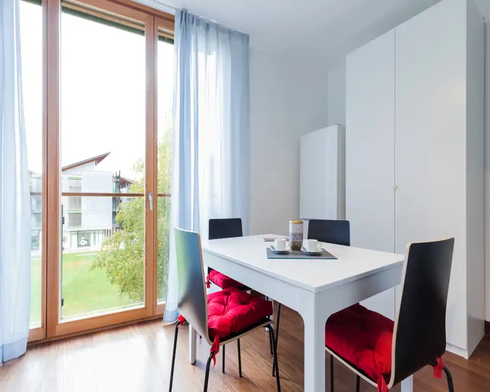 Bright, modern dining corner with a white table, four chairs with red seat cushions and large floor-to-ceiling windows that let in abundant natural light.