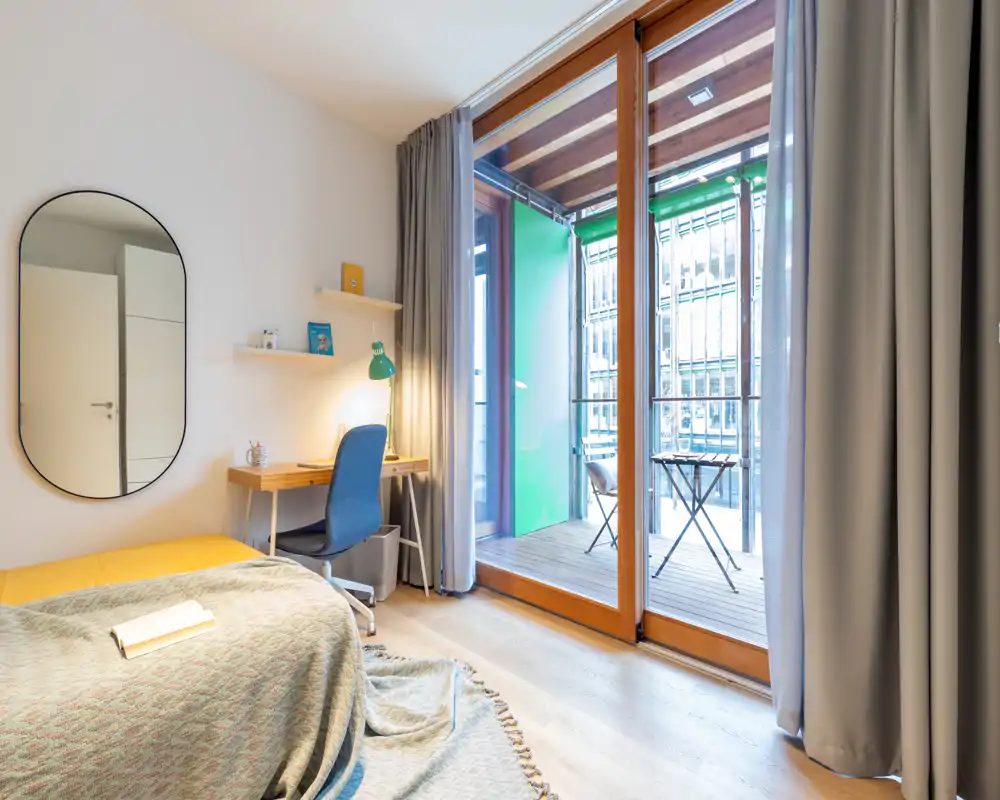 Sunny, modern bedroom with a comfortable bed, bedside textiles and a small desk. Large glass doors open onto a furnished balcony, filling the room with natural light.