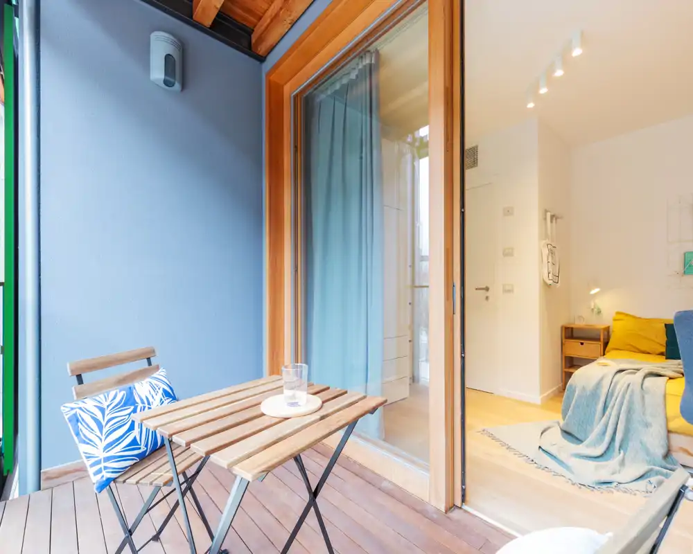 Bright, cozy bedroom seen from a small wooden balcony: a neatly made bed with pillows and a throw, soft natural light and warm wooden flooring create a welcoming atmosphere.