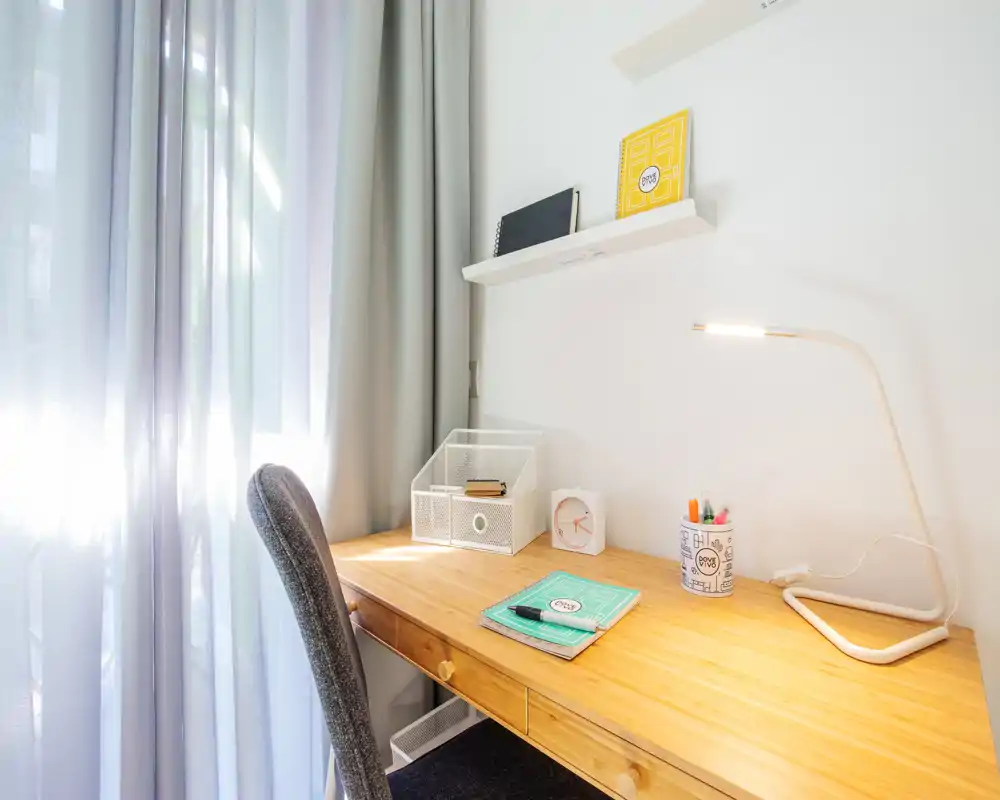Angolo studio luminoso e ordinato in camera, con scrivania in legno, sedia, lampada da tavolo e cancelleria vicino a una finestra con tenda leggera.