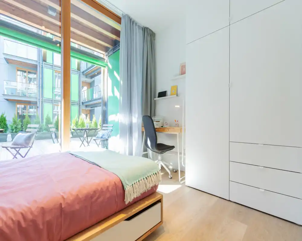 Camera da letto moderna e luminosa con letto comodo, ampia finestra a tutta altezza che si affaccia su una terrazza, area scrivania compatta e armadio a muro — perfetta per lavoro e relax.