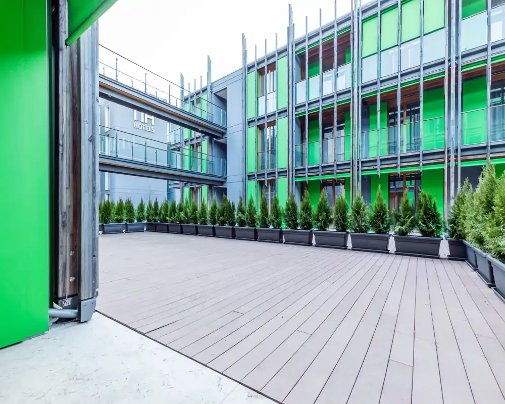 Amplia terraza exterior moderna con suelo efecto madera y una fila de macetas, enmarcada por una fachada contemporánea — ideal para descanso comunitario o eventos.