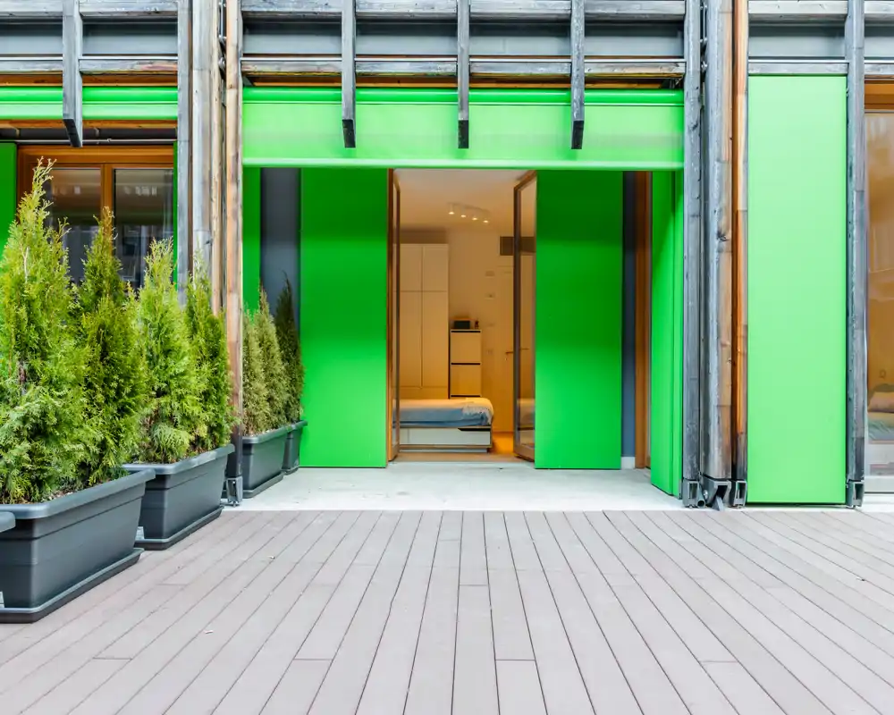 Dormitorio moderno visible a través de una abertura verde tipo escaparate; cama bien hecha, mobiliario minimalista e iluminación interior brillante. Buena opción para estancias urbanas compactas con acceso directo al exterior.