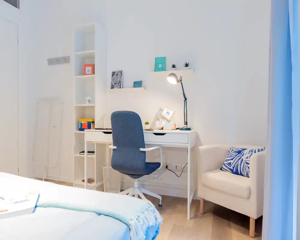 Rincón de dormitorio luminoso y moderno con cama visible en primer plano, escritorio blanco con silla giratoria azul y un sillón acogedor — ideal para trabajar y descansar.