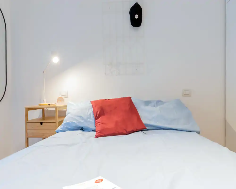 Dormitorio luminoso y minimalista con una cama doble bien hecha, ropa de cama azul claro y un cojín rojo como acento; mesita con lámpara y decoración de pared sencilla.