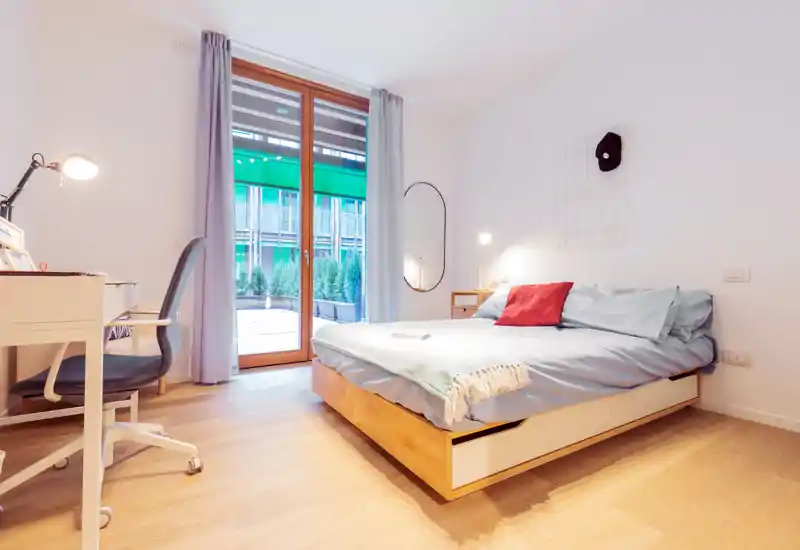 Dormitorio moderno y luminoso con cama baja de madera, ropa de cama azul y cojín rojo como toque de color. Gran puerta de cristal al exterior y un escritorio con silla que ofrece un espacio de trabajo funcional.