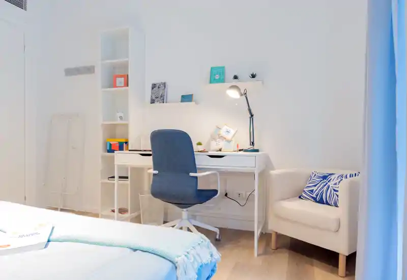Rincón de dormitorio luminoso y moderno con cama visible en primer plano, escritorio blanco con silla giratoria azul y un sillón acogedor — ideal para trabajar y descansar.