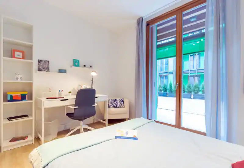 Habitación luminosa y bien decorada con cama en primer plano, escritorio funcional y estanterías, y una gran puerta de vidrio que da a una terraza exterior — perfecta para trabajar y relajarse.