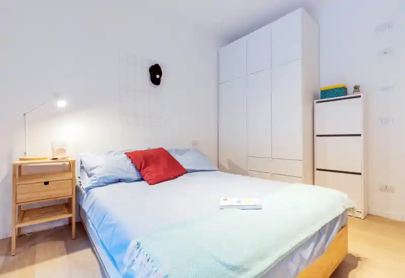 Dormitorio luminoso y bien presentado con cama doble de ropa de cama azul claro, cojín rojo de acento, mesita de madera con lámpara y armario alto blanco — ideal para anuncios de alquiler.