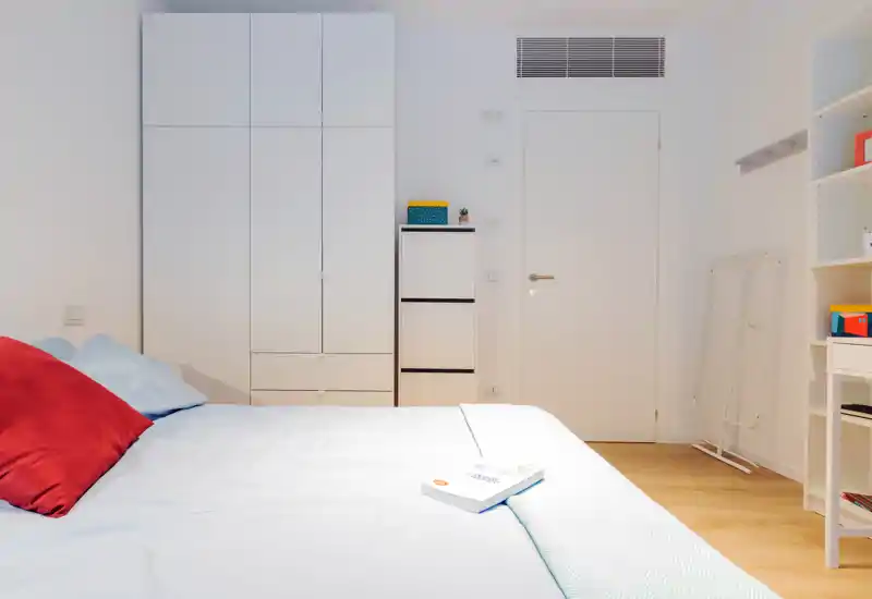 Dormitorio luminoso y minimalista con una cama grande, ropa de cama ordenada y armario blanco empotrado. La estancia está bien iluminada y despejada, perfecta para fotos de anuncio que destaquen espacio y confort.