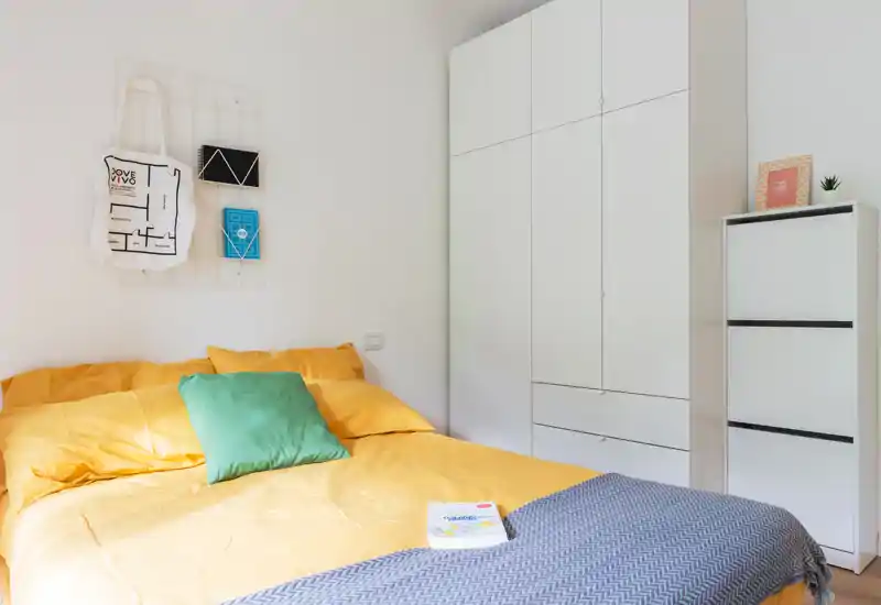 Dormitorio luminoso y ordenado con cama doble con ropa de cama amarilla, cojín verde y armarios empotrados blancos—moderno y bien iluminado.